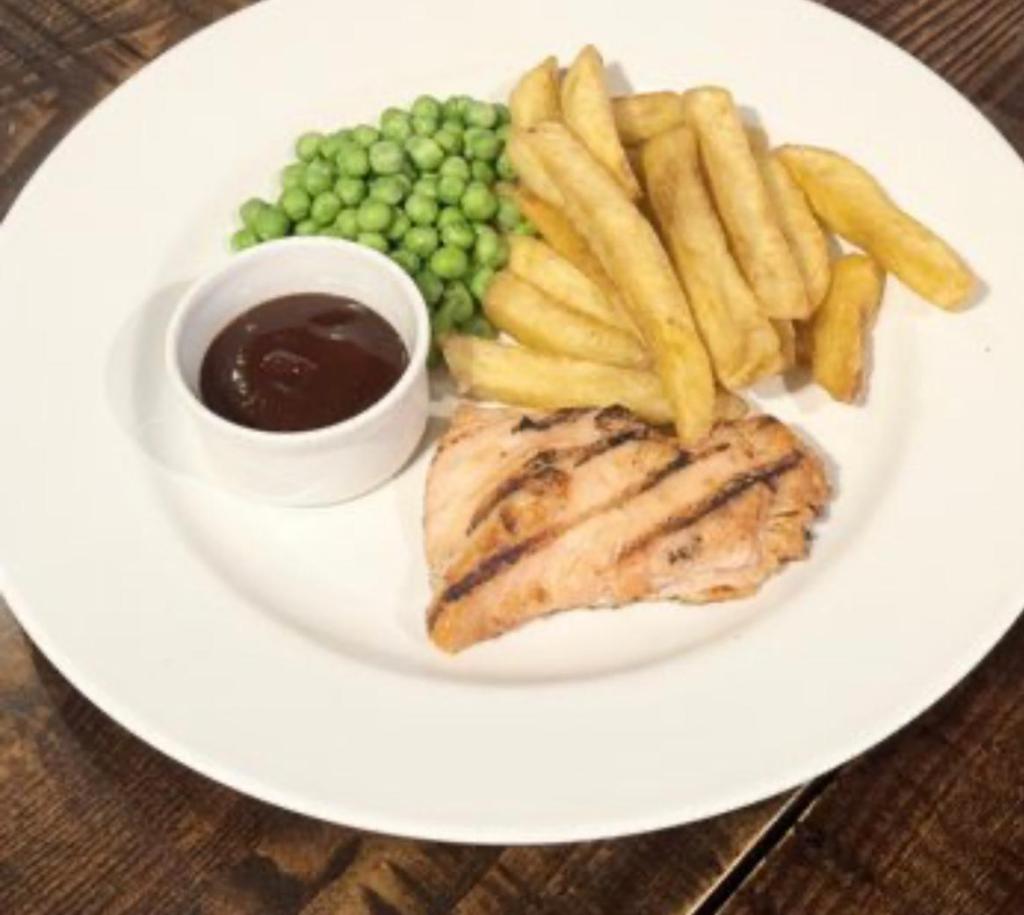 A plate full of chargrilled chicken breast with chips and peas on a wooden table. A pot of barbecue sauce sits besides the peas. 