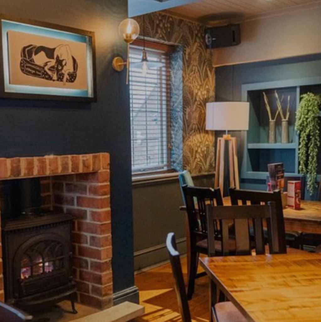 Pub interior, cosy looking space with a window allowing in natural light, tables and chairs are gathered around a fireplace, ready for diners.  