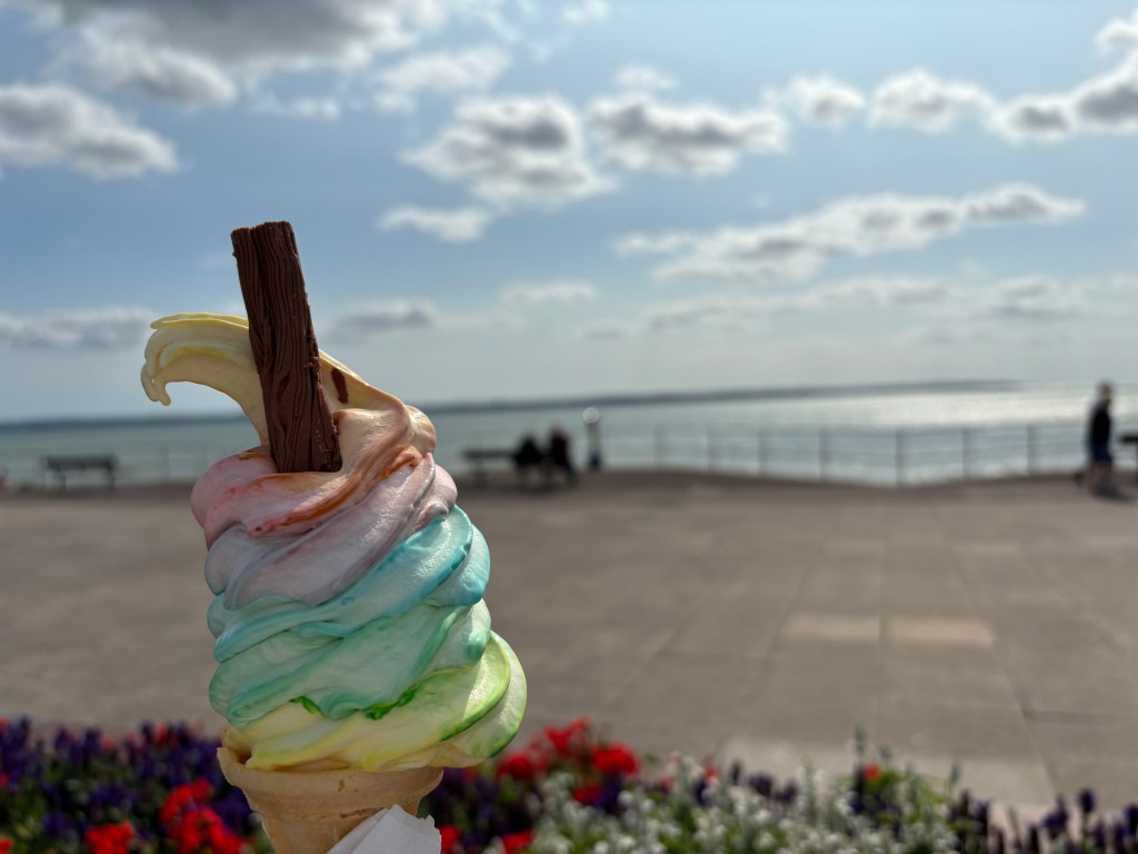 A colourful soft-whipped ice cream with a flake held in in front of a waterfront promenade with the Solent glistening behind.