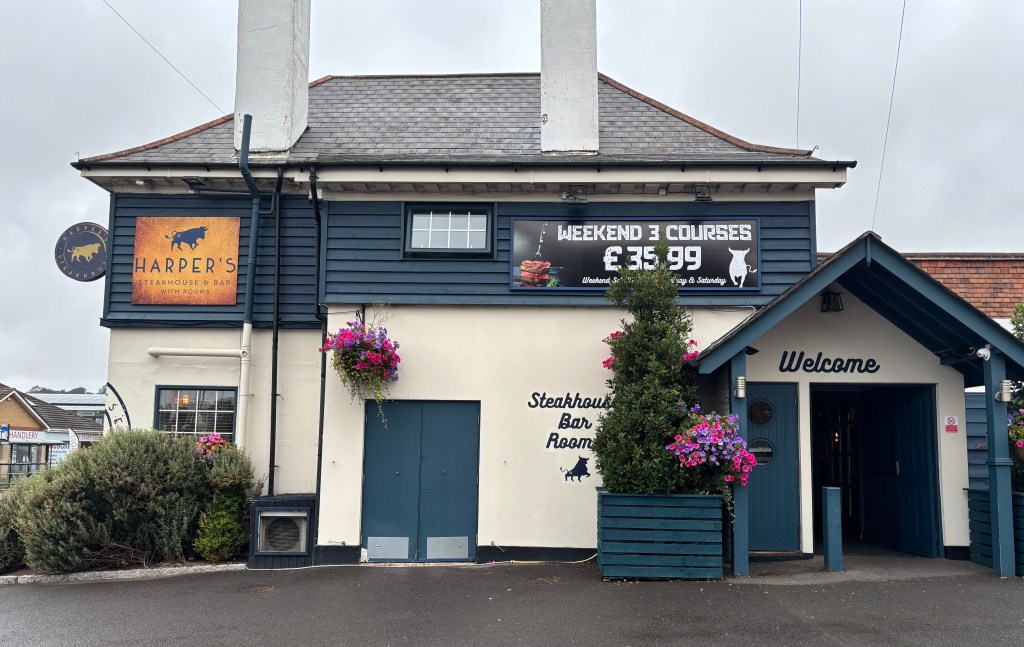 Harper's Steakhouse and Bar beneath a cloudy sky. Flower baskets hang outside, it looks classy with white paint and blue wood pannelling.