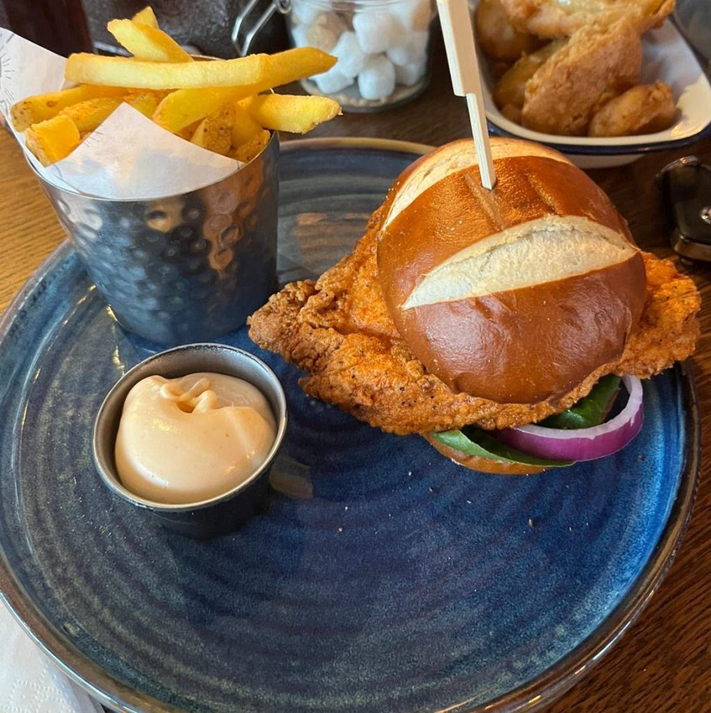 A buttermilk chicken burger with lettuce chunky onion in a pretzel burger bun. A pot of spiced mayo on the side, beside skin-on fry chips.