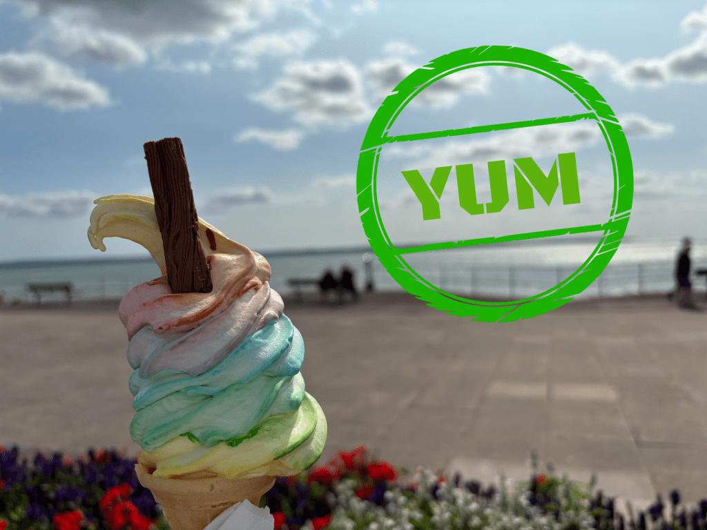 A colourful soft-whipped ice cream with a flake held in in front of a waterfront promenade with the Solent glistening behind. A large green, circular stamp saying Yum gives it special mention approval.