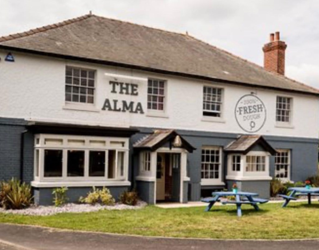 The front of The Alma Inn is painted white and grey. Picnic tables set up on the grass out front. Painting on the white section reads 100% fresh dough.